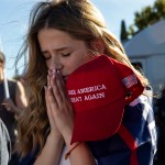 A young white woman prays with eyes closed while holding a MAGA red hat.