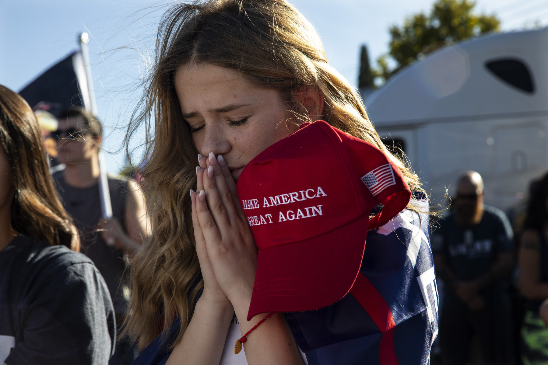A young white woman prays with eyes closed while holding a MAGA red hat.