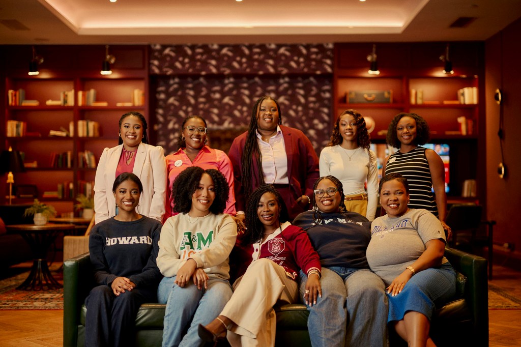 A group of Black women pose together in a warmly lit lounge, seated and standing in two rows, smiling at the camera.
