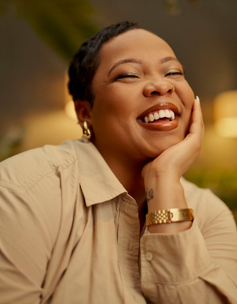 A close-up portrait of a woman smiling wearing a light-colored shirt and gold jewelry.