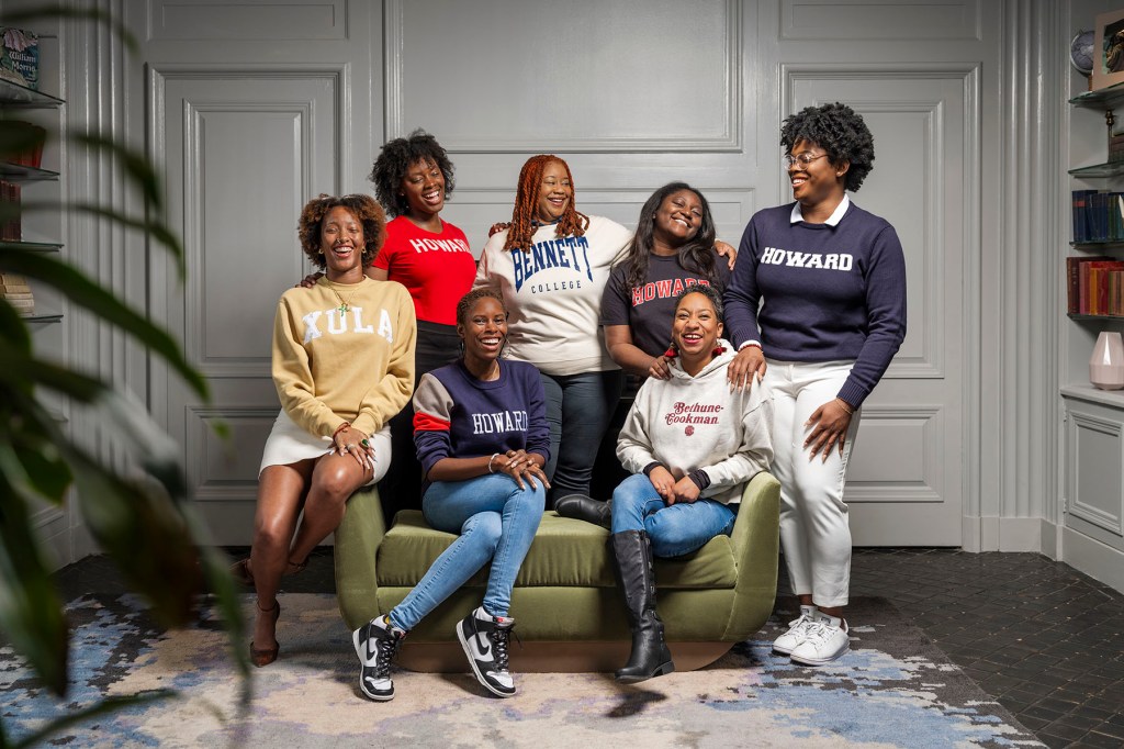 Eight Black women pose together in a studio setting, some seated and some standing, wearing sweatshirts representing historically Black colleges and universities.