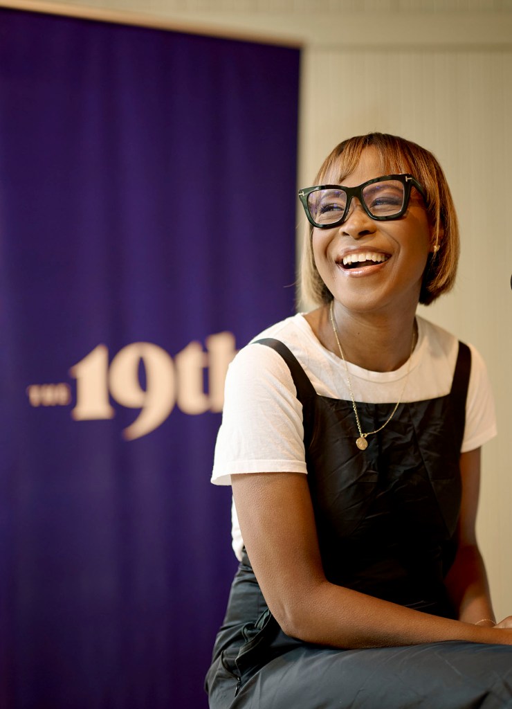 A Black woman wearing glasses laughs while seated indoors, with a purple banner reading “the 19th” blurred in the background.