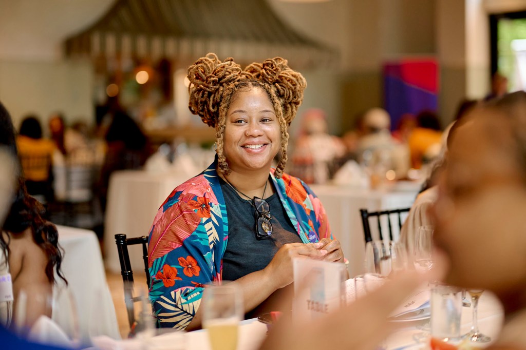 A woman with locs styled in two buns smiles while seated at a table during a group meal, with other attendees blurred around her.