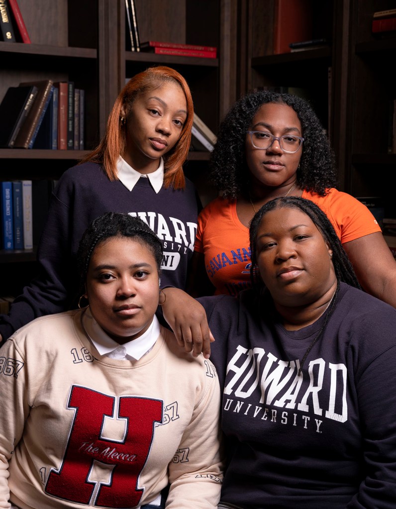 Four Black women pose closely together indoors in front of bookshelves looking directly at the camera.