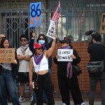 A group of people carrying signs and U.S. flags stand along a wall.