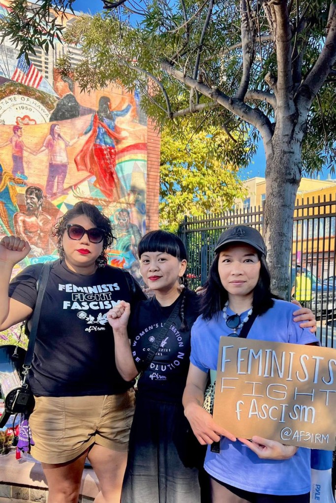 Three people stand together looking into the camera with raised power fists and one holding a sign that says “Feminists Fight Fascism.”