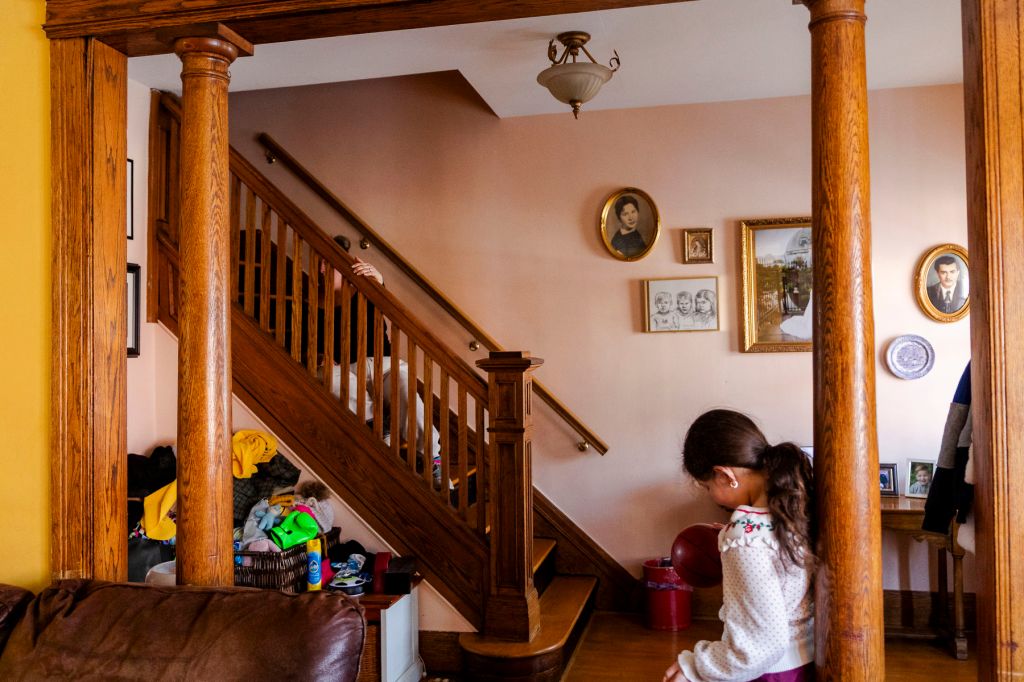 A child stands in a living room while her mother sits on the nearby stairwell grasping the banister.
