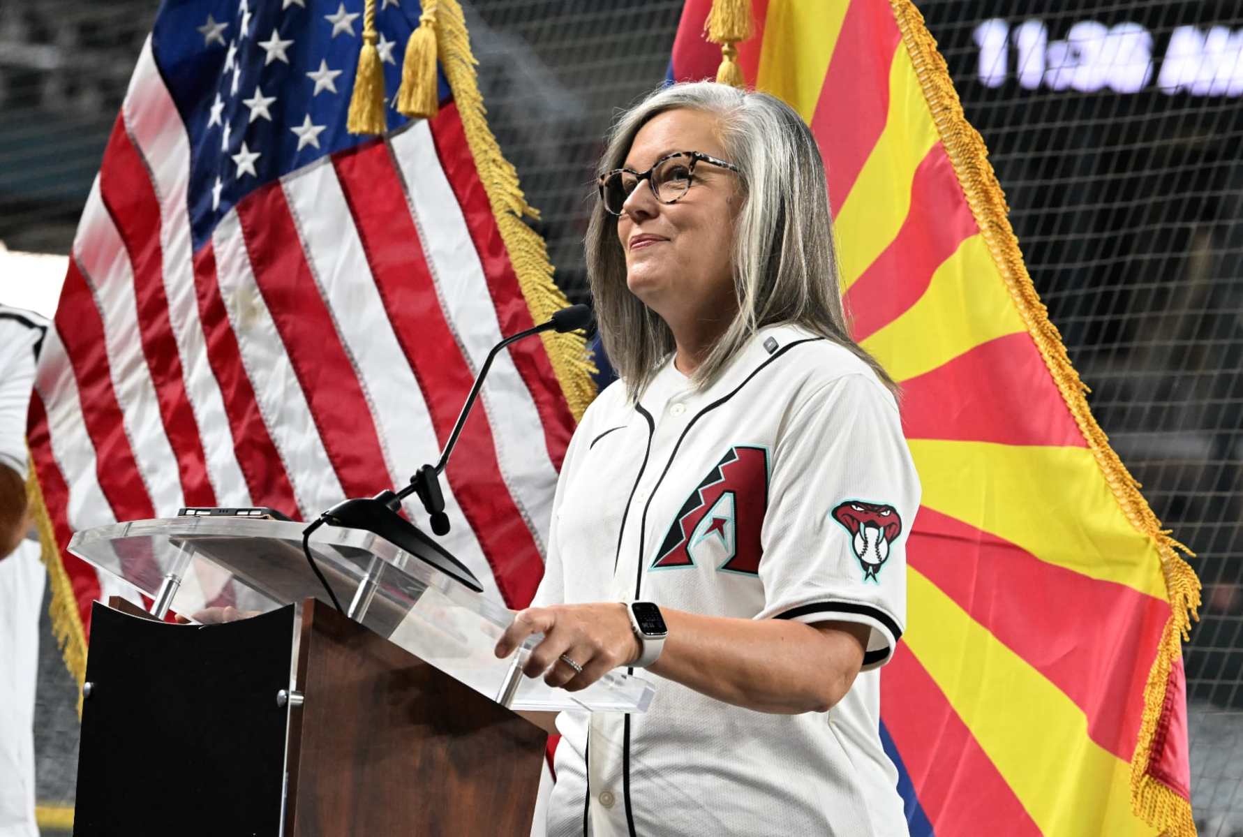 Katie Hobbs, in a Diamondbacks jersey, stands in front of Arizona and U.S. flags.