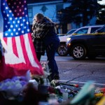 Two people hug while standing near a makeshift memorial for Renee Good, with flowers, candles and an American flag visible in the foreground and police tape and vehicles behind them.