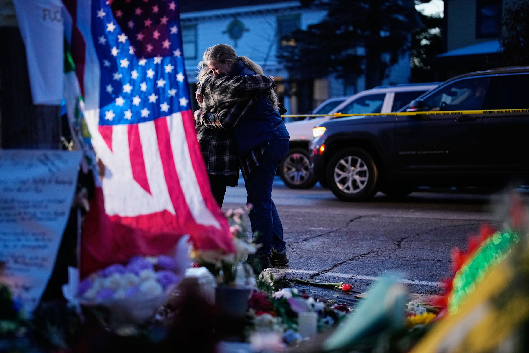 Two people hug while standing near a makeshift memorial for Renee Good, with flowers, candles and an American flag visible in the foreground and police tape and vehicles behind them.