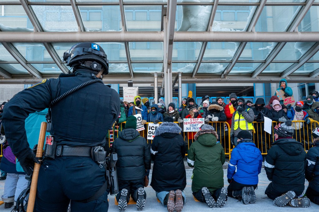 A line of demonstrators kneel behind a metal barricade during a protest at the Minneapolis–Saint Paul International Airport as a police officer stands in the foreground facing the crowd.