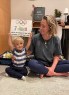 A woman sits on the floor beside a young child who holds a handmade sign reading “7 days until my mama is on the Olympic starting line,” with Olympic rings drawn above the text.