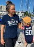 A woman wearing a “Squad 24 U.S. Sailing Team” shirt stands beside a young child on a sailboat dock, with masts and boats visible in the background.