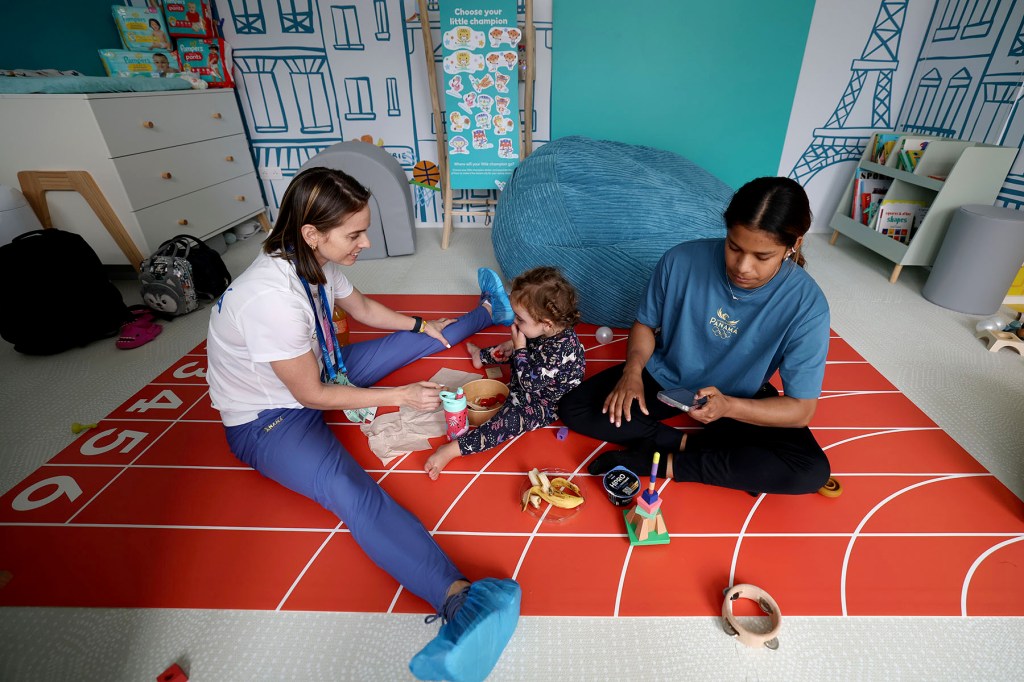 A woman and another adult sit on a playroom floor with a toddler, sharing food during a break in a childcare space decorated with toys, books, and a small indoor track mat.