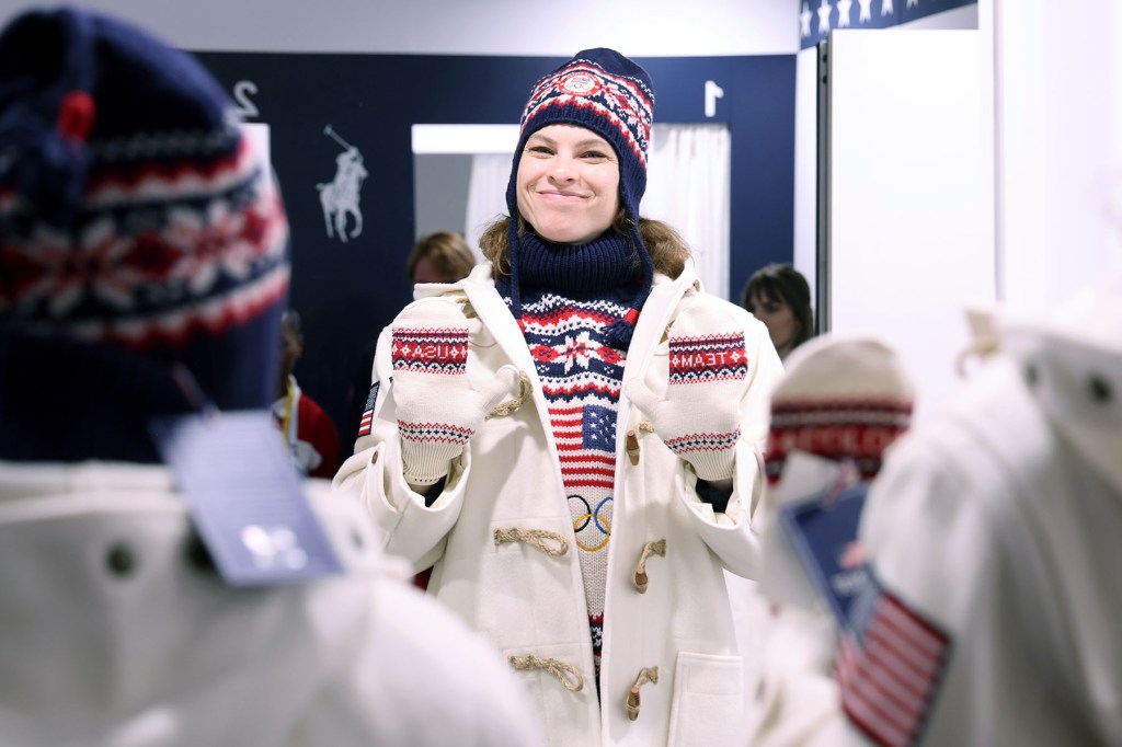 A winter sport athlete wearing a Team USA sweater, hat, and gloves smiles while standing indoors.