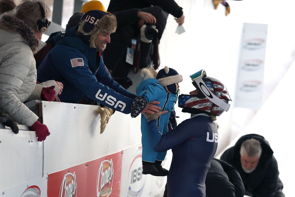 A man and woman lean over a barrier to pass a bundled baby to a helmeted Team USA athlete wearing a racing suit at an ice track.