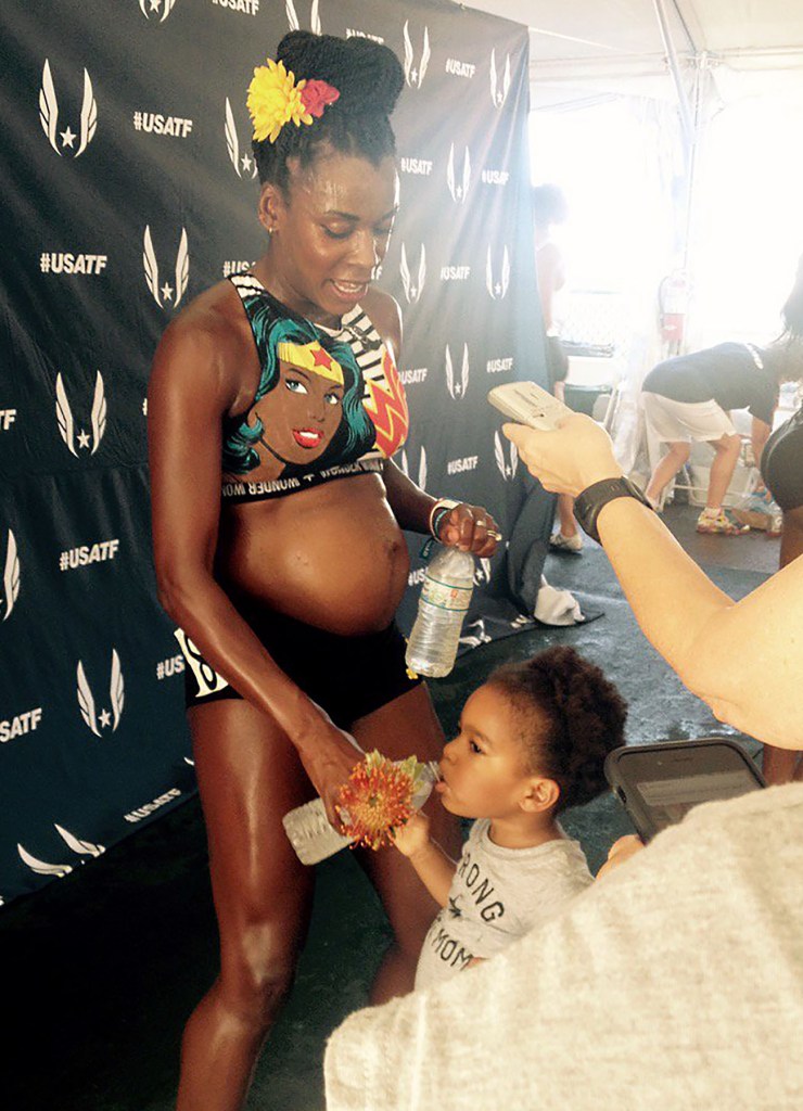 A pregnant track athlete stands holding a water bottle while a small child drinks from another bottle beside her, with a backdrop displaying “USATF” logos behind them.