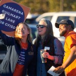Three people wearing campaign shirts smile as they take a selfie at an outdoor campaign event. One person holds a campaign sign reading “Camarena Guilmart & Lane for CFISD,” while cars are visible in the background.