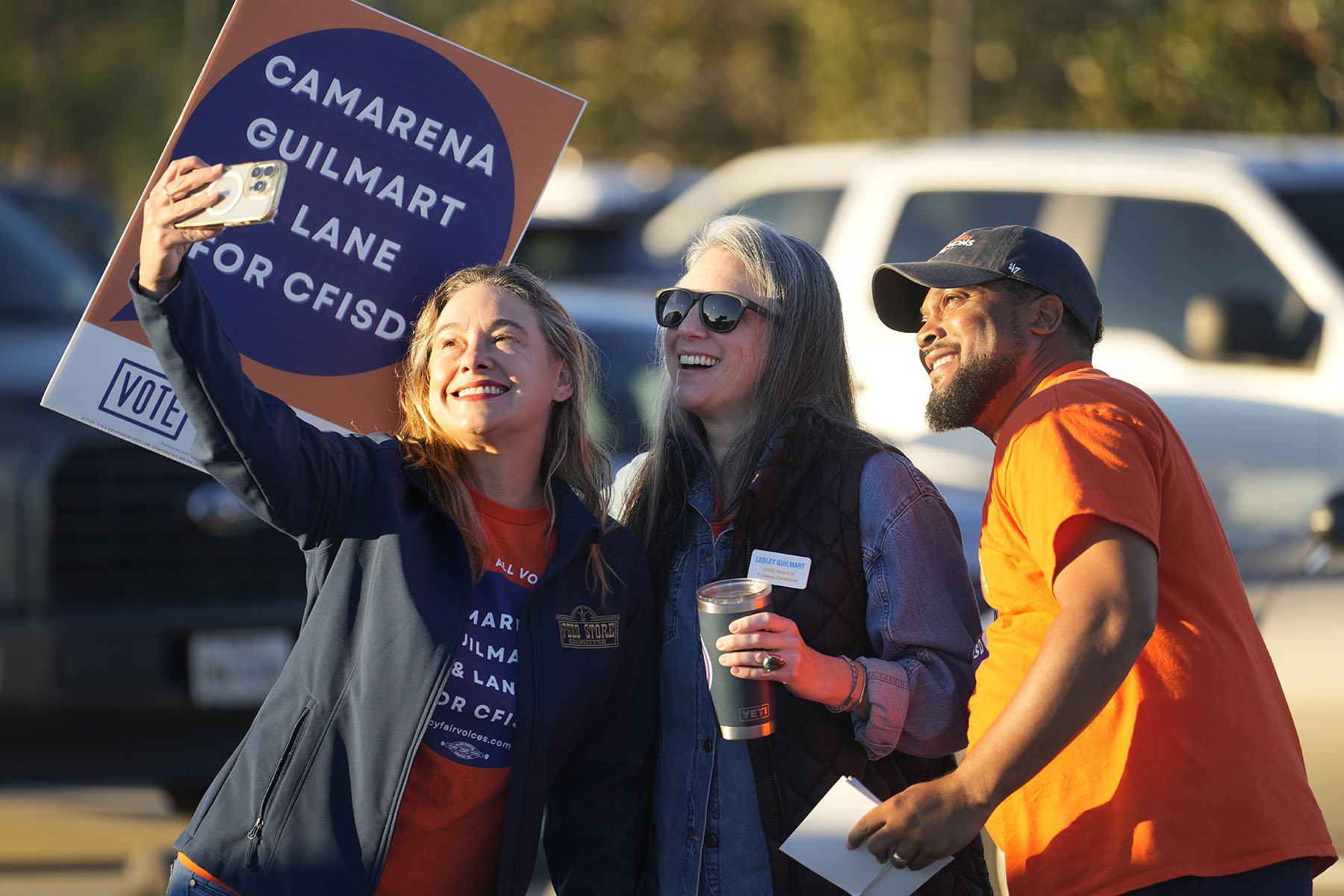 Three people wearing campaign shirts smile as they take a selfie at an outdoor campaign event. One person holds a campaign sign reading “Camarena Guilmart & Lane for CFISD,” while cars are visible in the background.