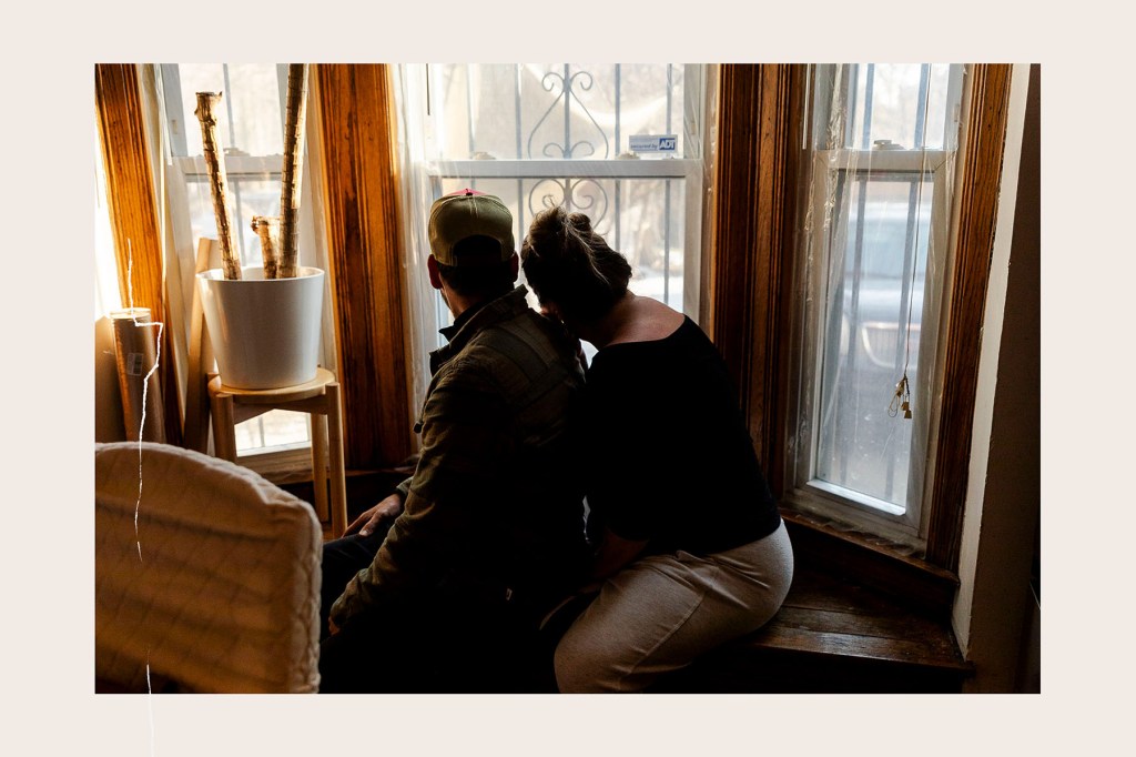 A man and woman sit close together on a wooden window bench, looking out through tall windows with afternoon light coming in. A potted plant on a small stand sits beside them.