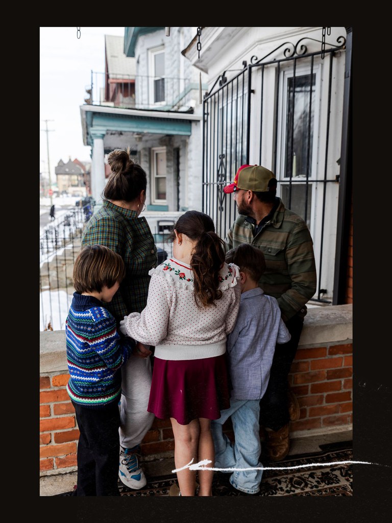 A family of five stands on a front porch behind a low brick wall, looking out toward the street. Two adults stand with three children clustered between them.
