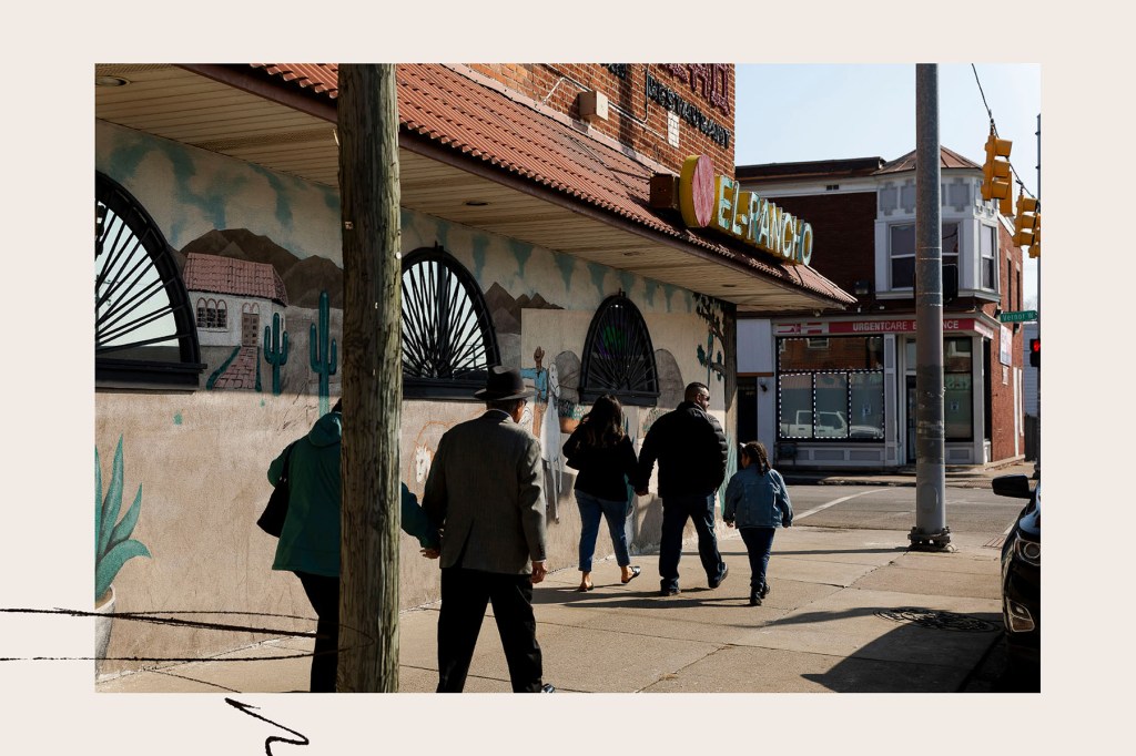 Several people walk along a sidewalk beside a building painted with a desert mural. A sign reading “El Rancho” hangs above the corner, and traffic lights stand at the intersection ahead.