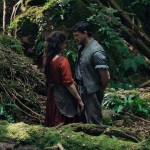 A woman and a man stand facing each other in a dense, mossy forest. The woman wears a long red dress and an apron, and the man wears rolled-sleeve work clothes with a vest. They appear to be speaking quietly while standing among fallen branches, ferns and green undergrowth.