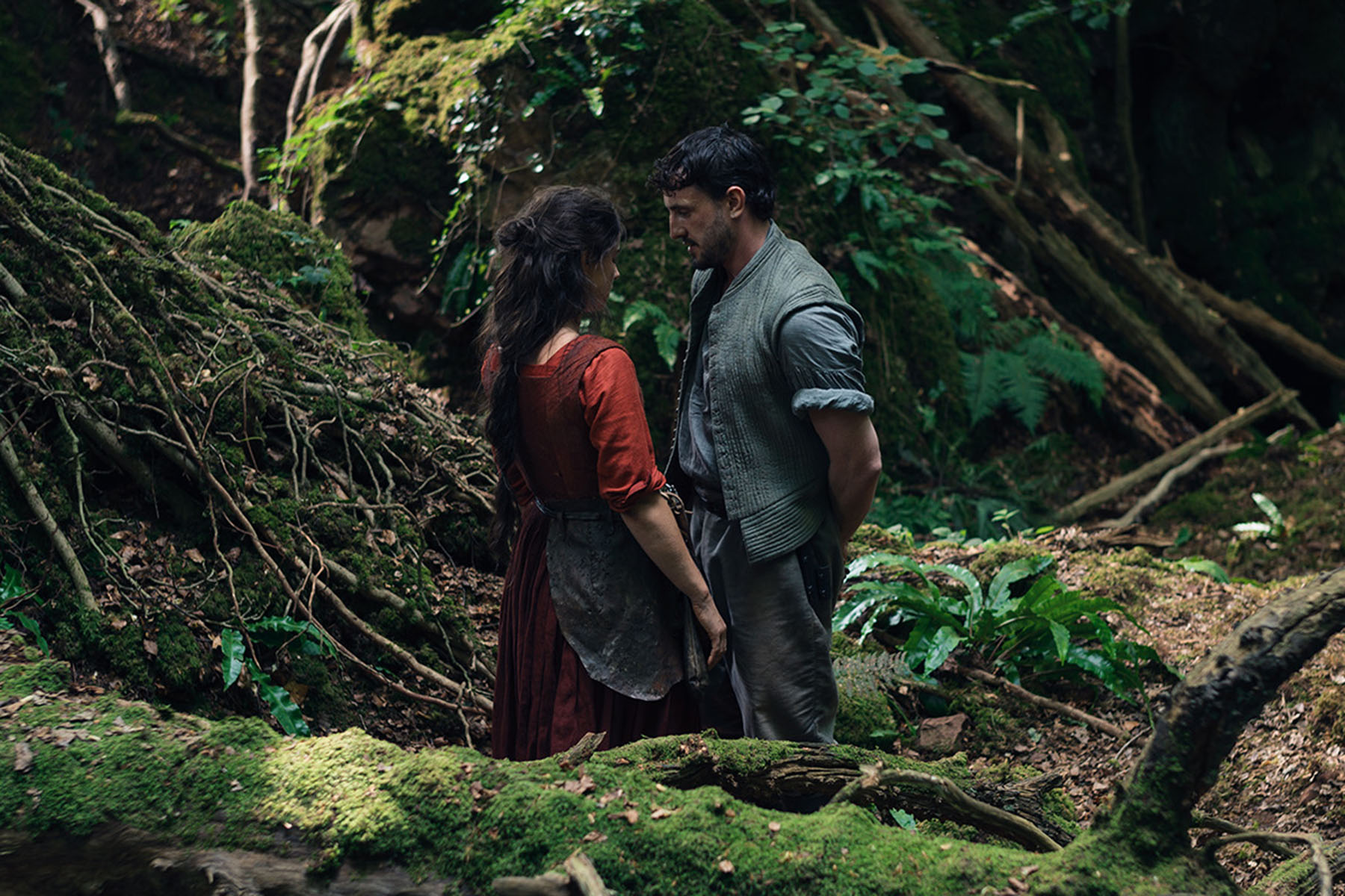 A woman and a man stand facing each other in a dense, mossy forest. The woman wears a long red dress and an apron, and the man wears rolled-sleeve work clothes with a vest. They appear to be speaking quietly while standing among fallen branches, ferns and green undergrowth.