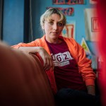 Kat Abughazaleh, in an orange hoodie and red T-shirt, sits on a couch indoors, looking directly at the camera, with colorful posters and a blue wall behind her.