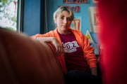Kat Abughazaleh, in an orange hoodie and red T-shirt, sits on a couch indoors, looking directly at the camera, with colorful posters and a blue wall behind her.