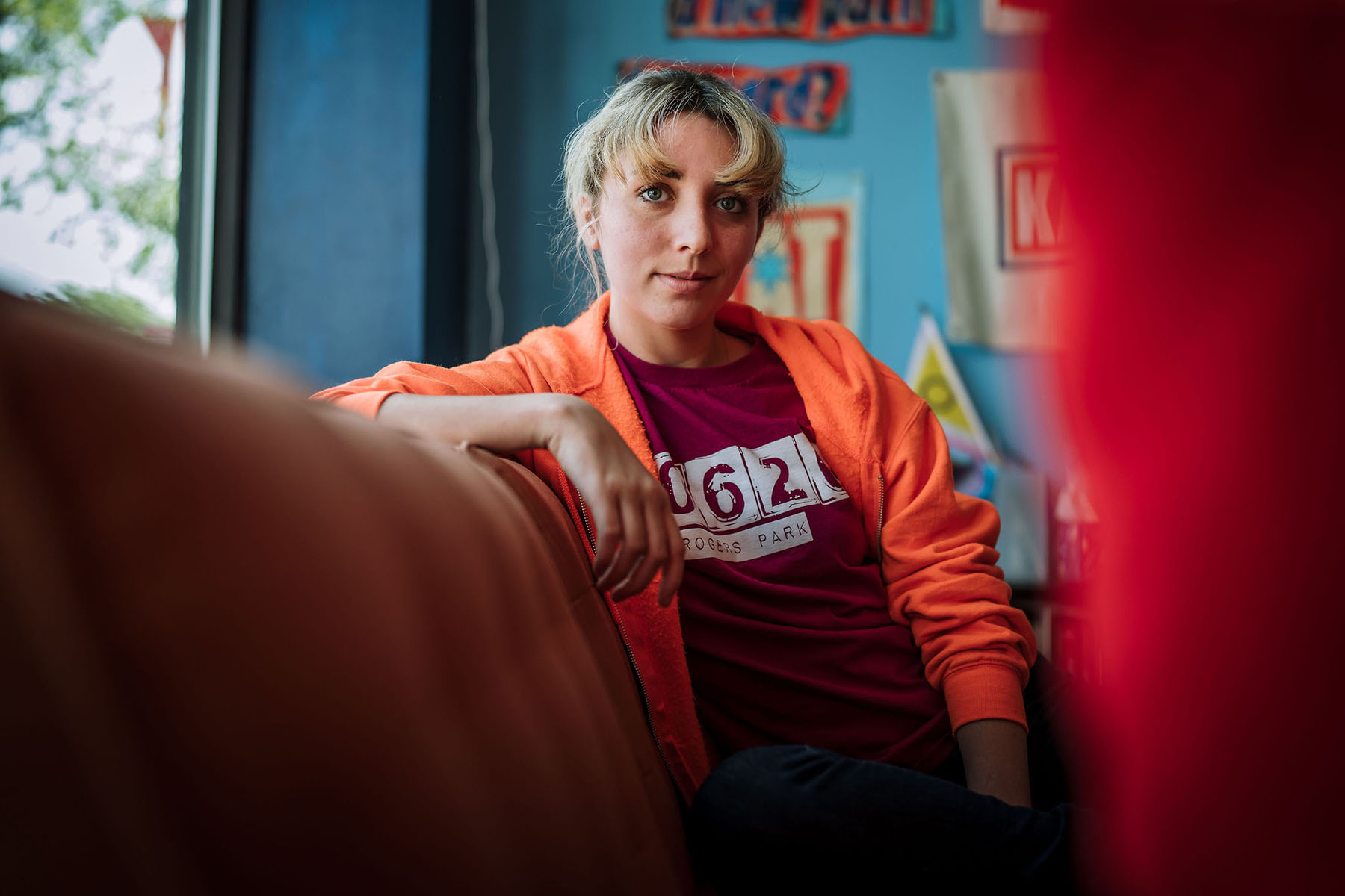 Kat Abughazaleh, in an orange hoodie and red T-shirt, sits on a couch indoors, looking directly at the camera, with colorful posters and a blue wall behind her.