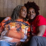 Two women sit close together on a couch, leaning against each other while facing the camera.