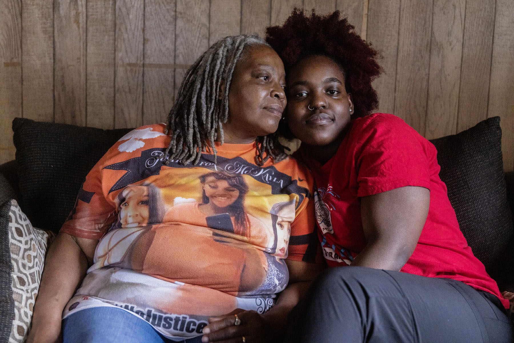 Two women sit close together on a couch, leaning against each other while facing the camera.