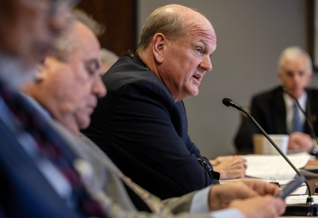 A group of men sit at a table during meeting with one man speaking into a microphone while others sit beside him.