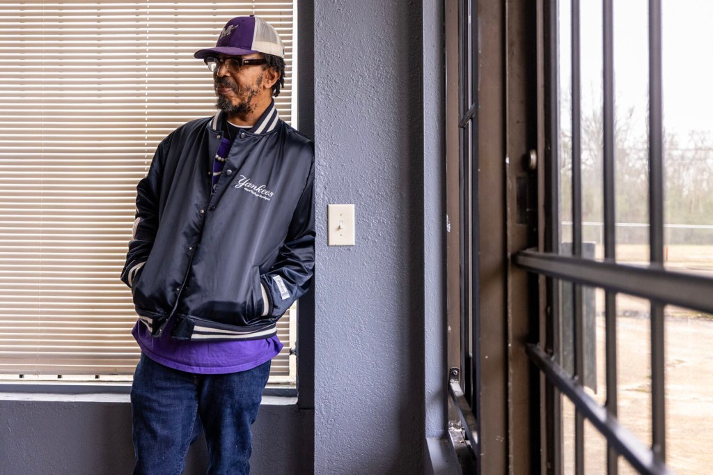 A man wearing a baseball cap and a dark jacket stands with his hands in his pockets beside a window inside a building.