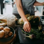 A woman arranges broccolini and aubergines on a slate at counter in a kitchen.