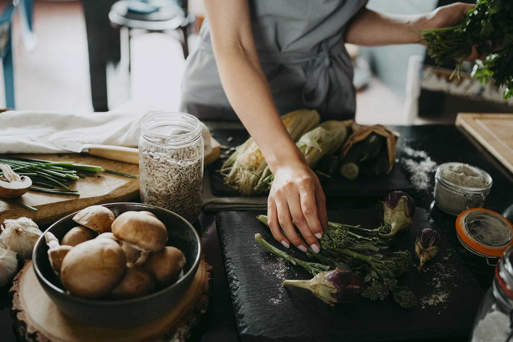 A woman arranges broccolini and aubergines on a slate at counter in a kitchen.