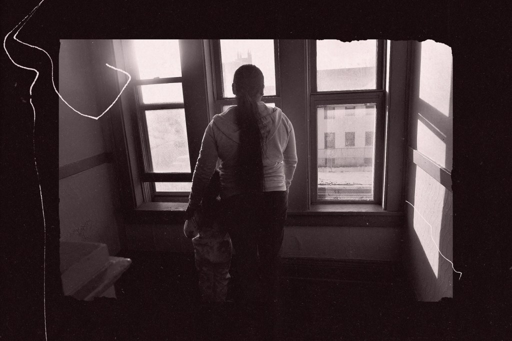 A person with a long ponytail stands at a window holding a small child, both facing outside in a dimly lit interior space.