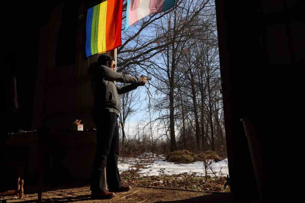 A person stands in the doorway of a structure aiming a handgun outdoors, with a rainbow flag hanging overhead and trees visible outside.