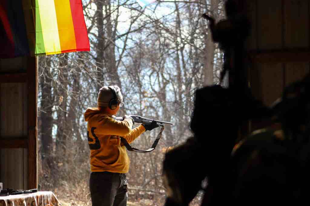 A person wearing hearing protection and a hoodie aims a shotgun outdoors from the entrance of a barn.