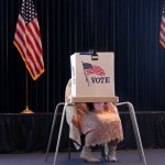 A woman wearing cowboy boots sits obscured behind a voting booth and flanked by tow U.S. flags.