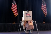 A woman wearing cowboy boots sits obscured behind a voting booth and flanked by tow U.S. flags.