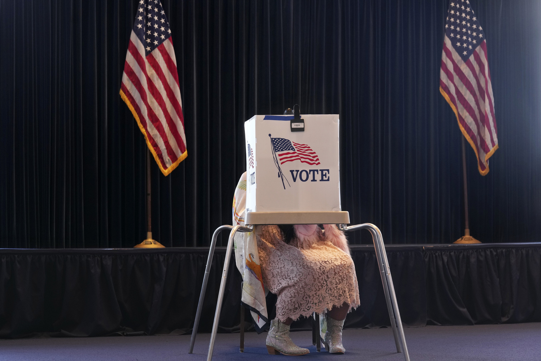 A woman wearing cowboy boots sits obscured behind a voting booth and flanked by tow U.S. flags.