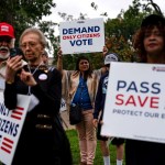 A group of people walk outdoors holding protest signs, including one that reads “Demand Only Citizens Vote” and another that says “Pass the SAVE Act - Protect Our Elections.”