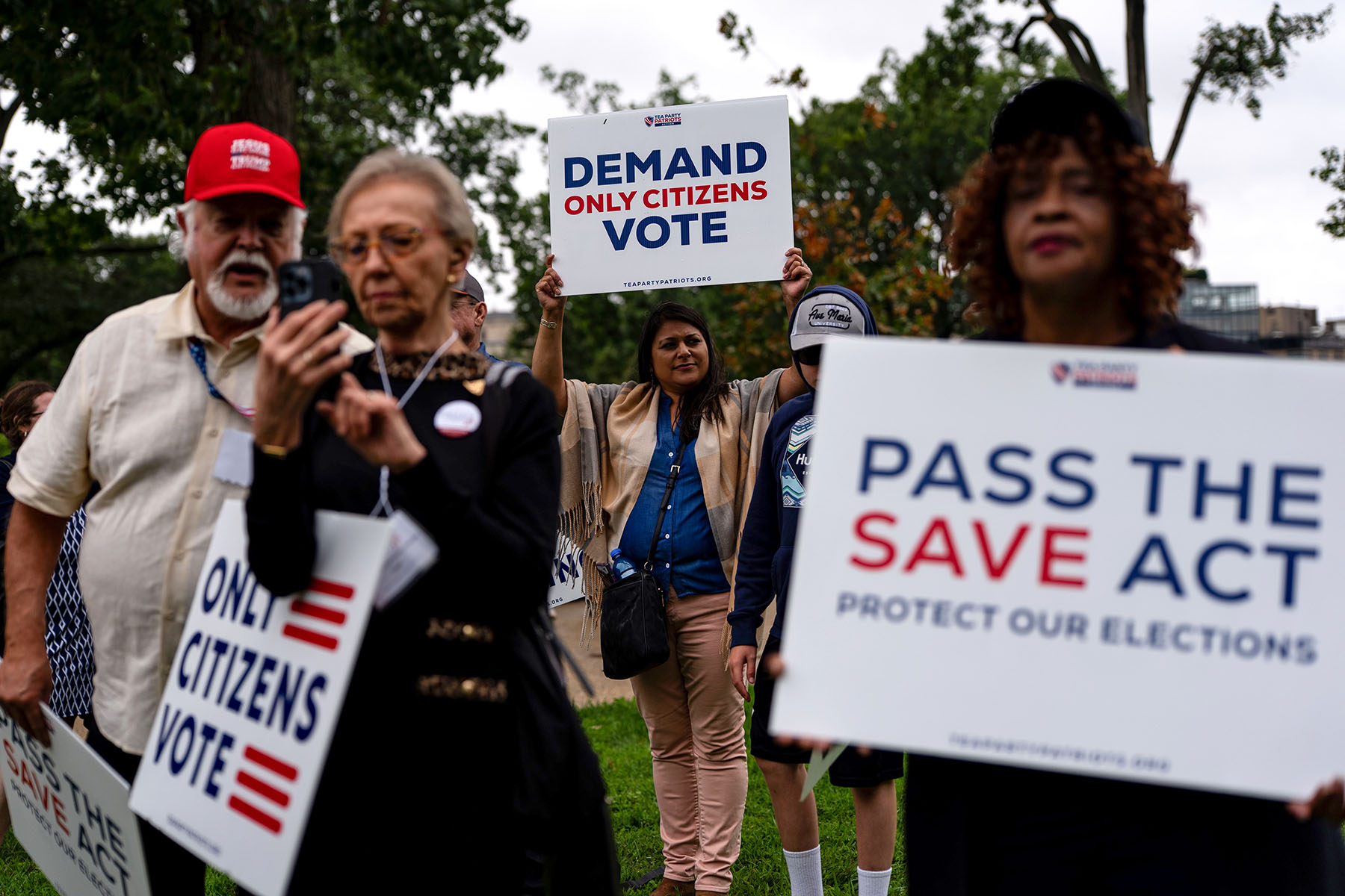 A group of people walk outdoors holding protest signs, including one that reads “Demand Only Citizens Vote” and another that says “Pass the SAVE Act - Protect Our Elections.”