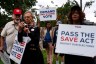 A group of people walk outdoors holding protest signs, including one that reads “Demand Only Citizens Vote” and another that says “Pass the SAVE Act - Protect Our Elections.”