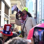 A protester wearing a neon yellow beanie speaks into a megaphone while standing above a dense crowd on a city street, with people around them holding signs and filming on their phones.