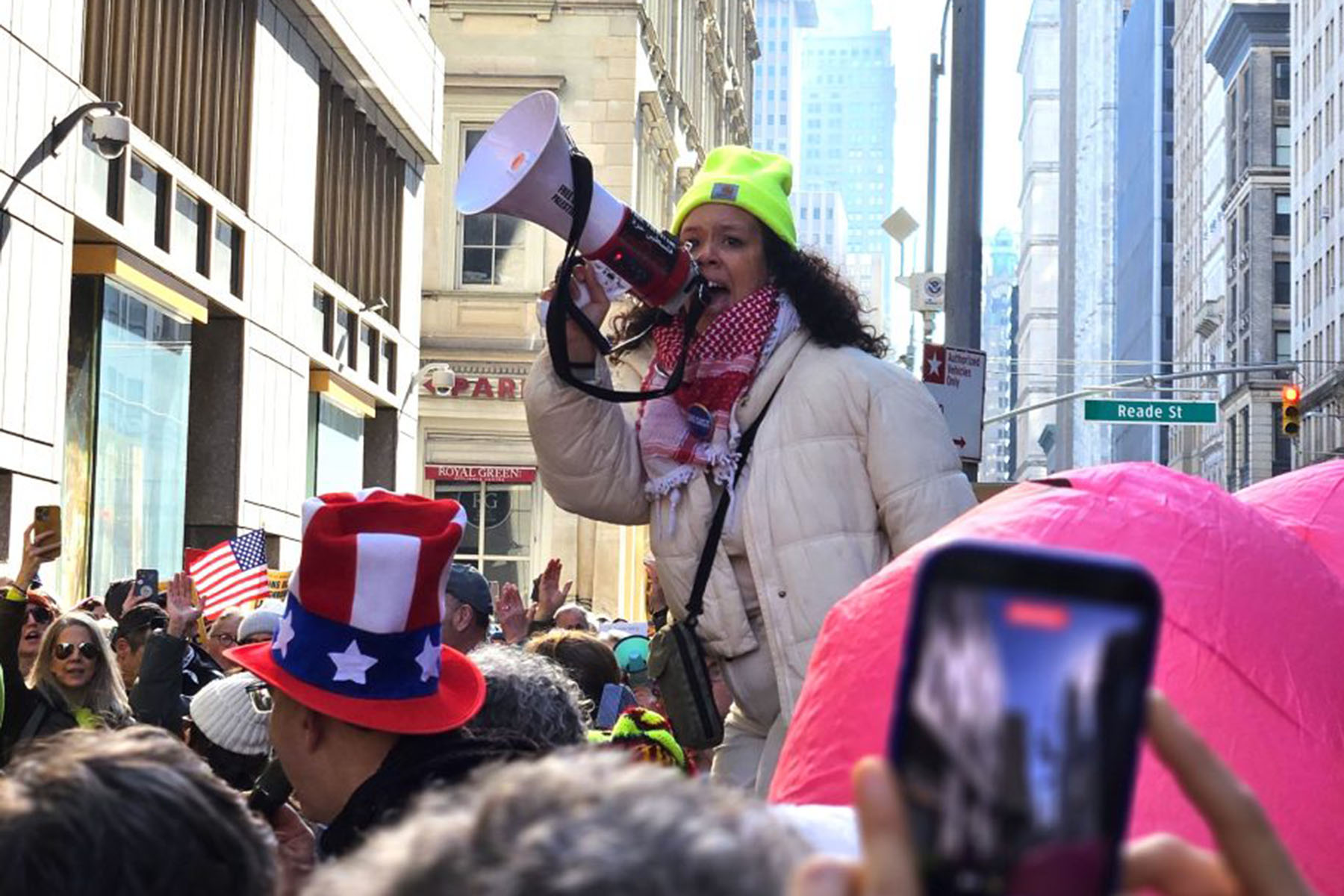 A protester wearing a neon yellow beanie speaks into a megaphone while standing above a dense crowd on a city street, with people around them holding signs and filming on their phones.