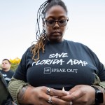 A woman wears a Save Florida ADAP t-shirt with her head bowed.