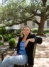 A woman sits outdoors on a wooden chair under large trees, with greenery and a building in the background.